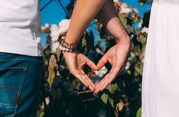 A couple show heart with hands in cotton field. Woman in white dress and man in blue jeans and white T-shirt. Girl and boy holding hands. Couple on valentines day. Clock and decoration on woman's hand