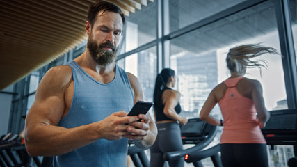Fototapeta na wymiar Muscular Heavyweight Champion Walks Through Gym, Uses Smartphone for Social Media and Conducting Business Affairs. In the Background Sports People Running on Treadmills