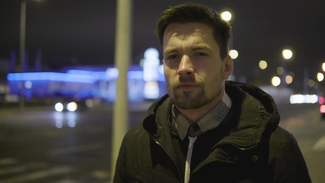 A young handsome man stands in a street in an urban area at night and looks into distance in thought - closeup