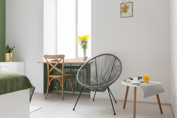a round armchair in a bright room with white walls, a cozy space. Wooden chair and flowers on the window. Interior in a modern minimalistic style.
