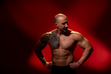 Fototapeta na wymiar A sports man with a tattoo posing on red background. Fitness trainer with naked torso, standing in the studio. Bodybuilder looking away