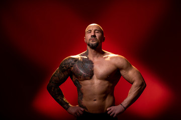 Fototapeta na wymiar Muscular man with tattoo and closed eyes posing on red background with naked torso, hands on the sides. Muscular body, studio shot