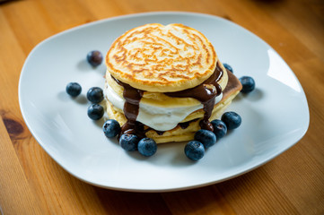 Pancakes with maple syrup and blueberries on a white plate with a wooden table as background or background. Sweet tasty breakfast or also as a delicious dessert. American pancakes with berries.