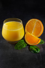 Glass with fresh orange juice, oranges and green leaves on a dark background.