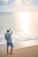 Asian man tourist wearing hat standing on tropical island beach enjoy looking at sunset. Relaxing holiday or vacation travel in summer season. Summertime trip concept © zephyr_p