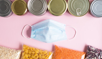 Corona virus special food supplies background concept. Medical mask protection in the middle of pasta, oatmeal, lentils, beans, canned food on pink background.