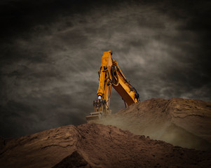 A yellow excavator bucket photographed from below behind a soil hill under a deamatic sky