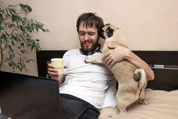 man lying on bed looking at laptop, his pug dog interfere, licking his face