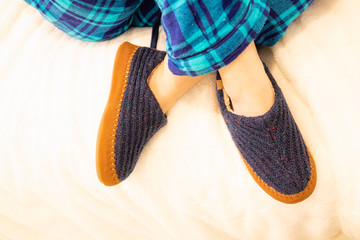 Person lounging in bed wearing blue pajamas and comfortable blue and tan slippers. Bright modern bedroom scene. 