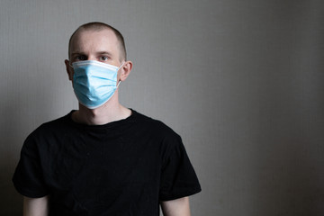 A guy in a medical mask and a black t-shirt is sitting on a light background