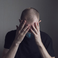 A guy in a medical mask and a black T-shirt is sitting on a light background, covering his eyes with his hands
