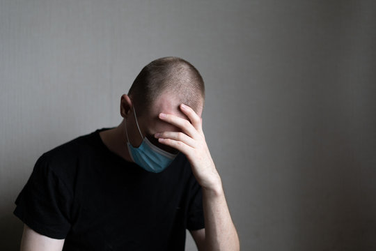 A guy in a medical mask and a black T-shirt is sitting on a light background, covering his eyes with his hand