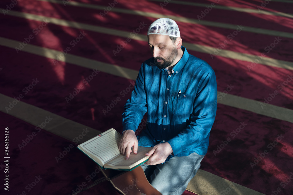 Wall mural Young Arabic Muslim man reading Koran and praying. Religious muslim man reading holy koran inside the mosque.