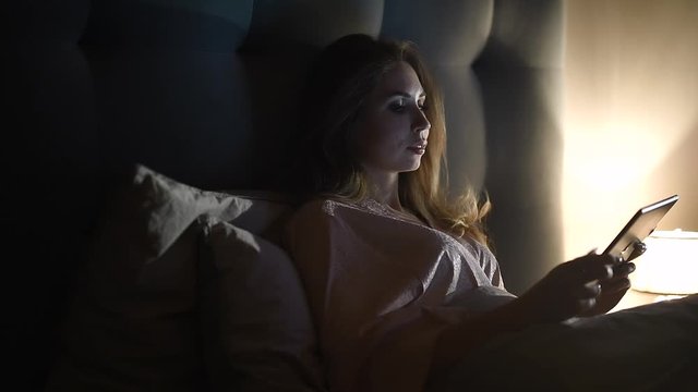 A young woman lying in bed and looking at the good news and photos from a trip on a tablet pc. Girl is reading an e-book in the bedroom.