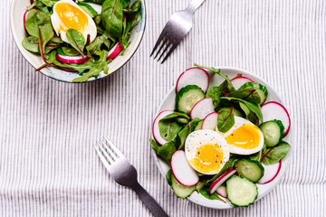 Radish, cucumber, herb mix and soft-boiled egg salad in two bowls on white tablecloth. Healthy nutrition. Selective focus