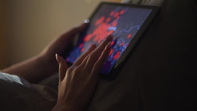 A young woman lying in bed and watching the news about the spread of the epidemic of coronavirus Covid-19. The girl in the bedroom leafs through the pandemic 2020 map on a tablet PC or smartphone.