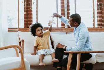 Happy family African American dad and son sitting on sofa playing with airplane wooden toy at home....