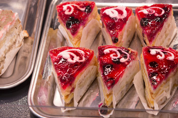 slices of fruit biscuit cake with jelly, wild berries, strawberries and whipped cream are cut and sold for sale in the pastry shop counter