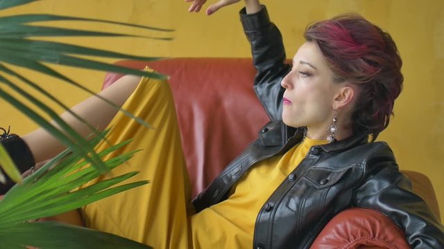 Close-up female portrait of glam rock style girl with dark pink hair and mohawk in yellow dress and black leather jacket chewing bubble gum sitting on armchair near the palm tree