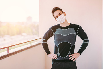 Young sporty man fitness coach in medical face mask listen music and train workout on balcony. Happy sports man in protective mask doing yoga exercises for stretching muscles. COVID-19 home quarantine