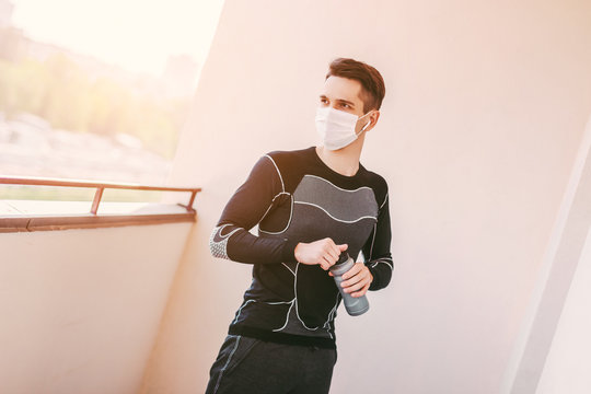 Young exhausted sports man in protective medical face mask holding bottle with water in hand while doing workout exercises on balcony. Tired thirsty fitness man drink water. COVID-19 home quarantine