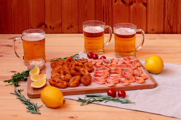 Fried squid rings and shrimps on cutting board and mugs of beer near. Selective focus.