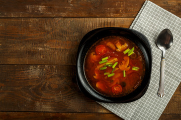 Black dish of italian tomato soup with potatoes and mushrooms on a wooden table on a napkin and a spoon.