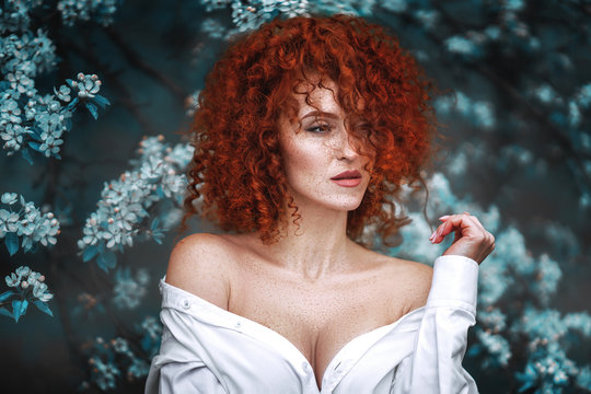 A red-haired girl with a very curly and voluminous hairdo in a shirt with bare shoulders looks into the distance. Her face and body with freckles. Art photo