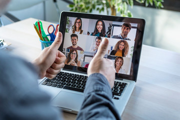 Male employee making a gesturing approval with thumb fingers while speak on video call with diverse colleagues on online briefing with laptop at home.