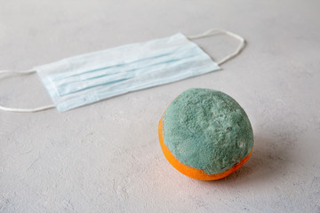 Rotten, spoiled, inedible orange with mold lies on a gray background. At the back is a medical mask that protects against coronovirus. Substandard product. Close up composition.