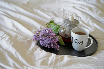 Tea pot and cup of tea with lilac flowers in bed