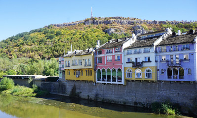 Four houses painted in violet, blues, yellow, pink and green on the shore of a river, at the base of a hill.