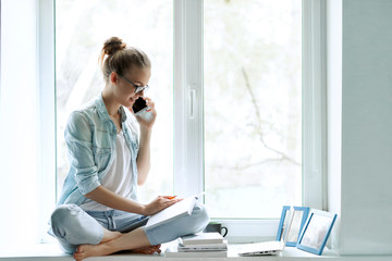 Young woman working from home office. A freelancer uses a laptop, phone, internet and notepad. Workplace on the windowsill. The concept of female business, career, communication, online learning