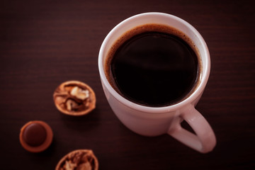 In a white deep mug is poured black coffee without milk, on the sides of the mug a little foam, in the background is a whole walnut and one sweet candy
