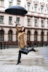 Outdoor photo of brunette lady jumping with black umbrella in rainy autumn day.Fashion street style portrait. wearing dark casual trousers, white sweater and creamy coat.Fashion concept.