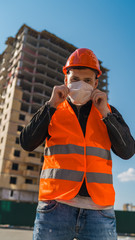 Man construction worker in overalls putting on medical mask on face on background of house under construction.