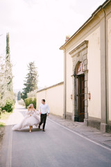 Fototapeta na wymiar Beautiful bride and groom running hand in hand outside of old villa in Italy, in Tuscany, near Florence.