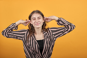 Cute Young Woman Light Brown In Striped Pink And Black Shirt On A Yellow Background, Happy Girl Smiles And Pulls Her Hands Behind Ears