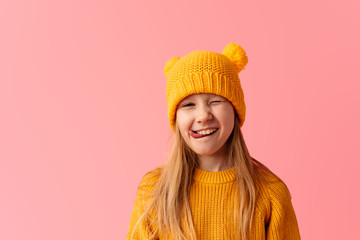 Happy little girl dressed for spring wearing yellow hat on pink background sticking out her tongue