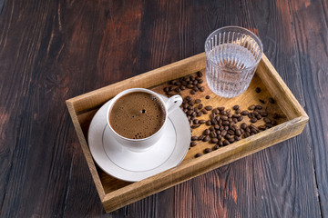 A Cup of coffee, coffee beans and glass of water in a wooden box on a dark wooden background.