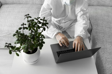 Man in protective white suit and medical mask is working from home on a sofa with laptop because of coronavirus epidemic. Remote work during pandemic. Stay home during COVID-19 quarantine concept.