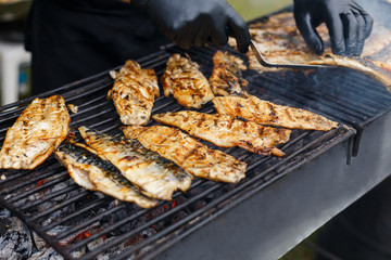 Cook in black suit remove hot fish from grates. A lot of mackerel lie on the grill. Catering food preparation for outdoor party. Barbeque for big company