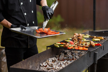 Cook in black suit remove hot pepper from grates. A lot of vegetables lie on the grill. Catering food preparation for outdoor party. Barbeque for big company