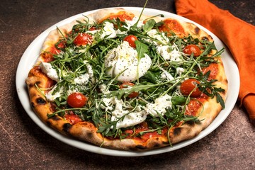 Pizza with mozzarella, tomato,arugula, thin dough, on plate, orange napkin. Brown background, top view