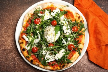 Pizza with mozzarella, tomato,arugula, thin dough, on plate, orange napkin. Brown background, top view