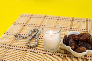 Ramadan Kareem Festival. Close up of dried dates fruit in bowl with candle and rosary on yellow background. Copy space.