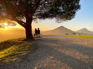 Silhouette of male-female couple sitting on bench under oak tree watching orange glow of sunset over mountains. Several other couples off in the distance