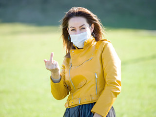 Beautiful woman in medical mask holding her middle finger up on nature. Coronovirus, pandemic, virus