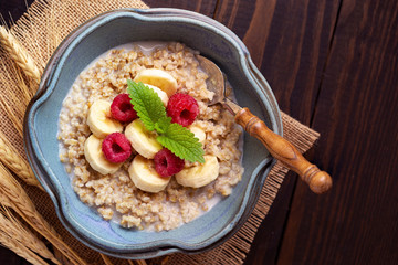 Overhead view of cooked steel cut oats topped with raspberries and bananas with a sprig of mint on a wooden surface with copy space