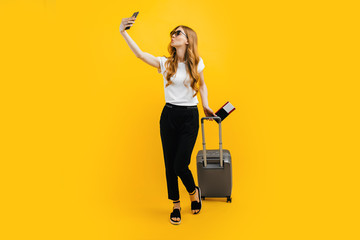 woman in sunglasses taking a selfie on a mobile phone, standing with a suitcase and a passport with tickets isolated on a yellow background.